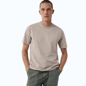 A man with short hair wears the HARTFORD Slub Cotton Crewneck T-shirt in Sand and green pants, standing with one hand in his pocket and looking to the side against a light background.