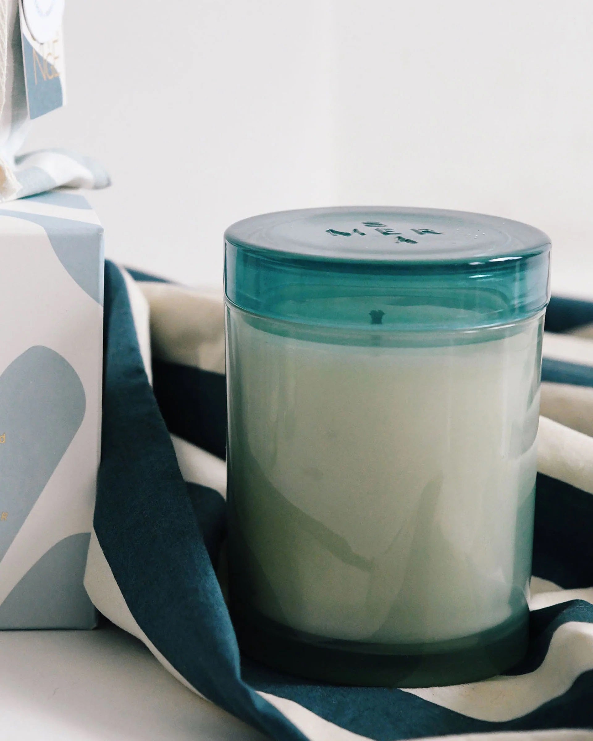 The MERSEA - SEA CHANGE JOLIE JAR CANDLE by MERSEA features white tea and sea salt-scented white wax in a cylindrical glass jar with a translucent blue lid, displayed against a white background.