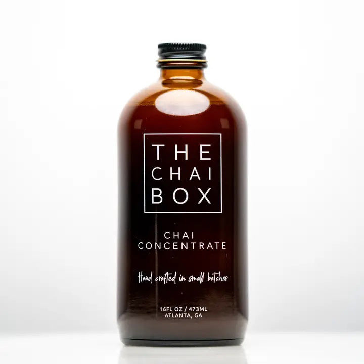 The Chai Box's brown glass bottle, prominently labeled with "THE CHAI BOX - CHAI CONCENTRATE," features text that reads "Hand crafted in small batches" and specifies a volume of 16 fl oz / 473 ml. Made with the finest spices, the label proudly displays "Atlanta, GA" at the bottom. The bottle is topped with a black screw cap.