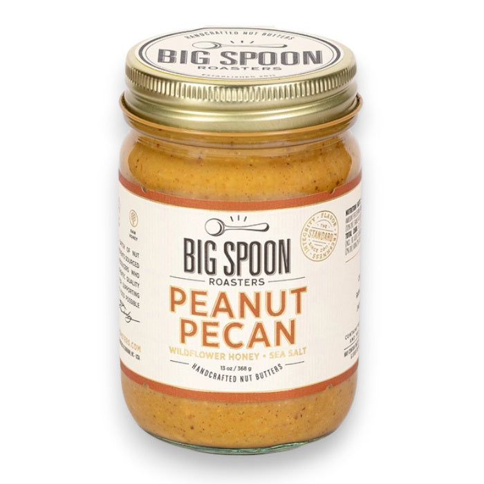 A jar of BIG SPOON ROASTERS Pecan Peanut Wildflower Honey Nut Butter with a gold lid. The label shows it contains wildflower honey, organic coconut oil, and sea salt. The jar is 13 oz or 369 g and is described as handcrafted nut butter from BIG SPOON ROASTERS.