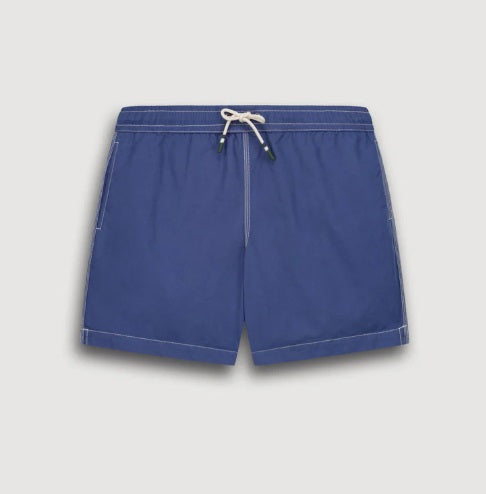 A shirtless person models the HARTFORD - NYLON SWIM TRUNKS WITH TRAVEL POUCH IN DENIM by HARTFORD against a plain light background, with tattoos visible on their arms and legs and one hand casually in a pocket.