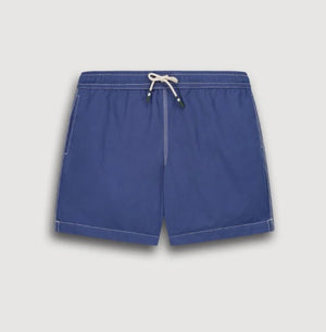 A shirtless person models the HARTFORD - NYLON SWIM TRUNKS WITH TRAVEL POUCH IN DENIM by HARTFORD against a plain light background, with tattoos visible on their arms and legs and one hand casually in a pocket.