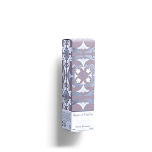 A tall, clear glass bottle of ROSE ET MARIUS’s A Stroll Under the Oratory Eau de Parfum 30ml, unisex with a light golden liquid and decorative floral-patterned square cap, is placed to the side on a white background.