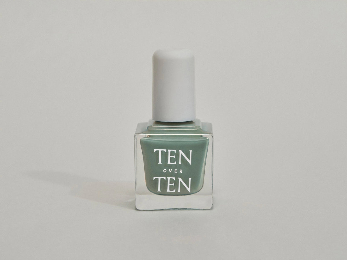 A glass bottle of TENOVERTEN - NAIL POLISH from the brand TEN OVER TEN, featuring a white cap and a dusty pink nail color. The front label displays "TEN OVER TEN" in white uppercase letters. This vegan, eight-free formula ensures both beauty and peace of mind. The background is plain and light-colored.