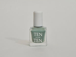 A glass bottle of TENOVERTEN - NAIL POLISH from the brand TEN OVER TEN, featuring a white cap and a dusty pink nail color. The front label displays "TEN OVER TEN" in white uppercase letters. This vegan, eight-free formula ensures both beauty and peace of mind. The background is plain and light-colored.