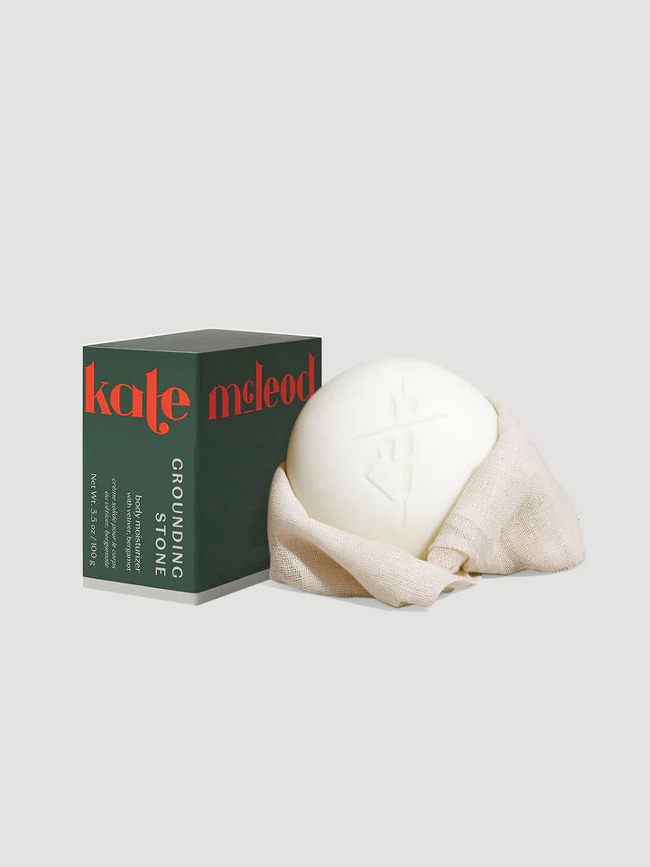 A white grounding stone wrapped in fabric sits beside a dark green box with "Kate McLeod" embossed in red. The box emits an earthy aroma, has details in white, and contains the Kate McLeod Grounding Stone Deep Moisturizing Lotion Bar Refill for skin nourishment.