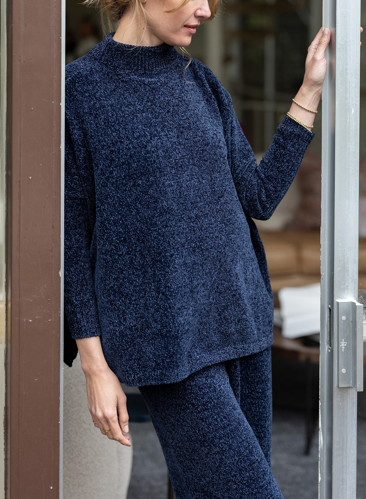 A woman stands in a doorway, wearing the MERSEA - CATALINA EVERSOFT CHENILLE SWEATER in relaxed dark blue. Her face is partially out of frame as she rests a hand on the door, effortlessly expressing cozy luxury.