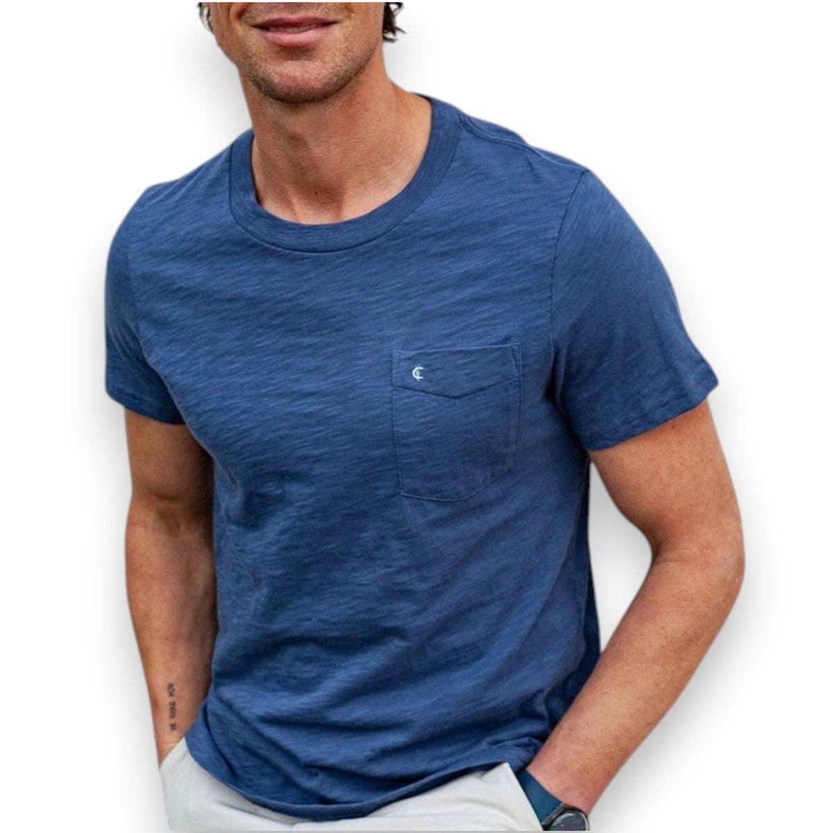 A man wearing the CRIQUET Slub Cotton Short Sleeve Pocket T-Shirt in Vintage Indigo stands against a textured white wall, paired with light-colored pants and one hand in his pocket. His face is partially out of frame.
