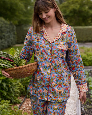 A woman with light brown hair sits against a white wall, wearing the PRINTFRESH - Bee Tapestry Long Pajama Set in Delphinium Blue. This organic cotton pajama set from PRINTFRESH features a colorful floral and bee pattern with vibrant red and yellow cuffs, collar, and pocket trim, beautifully blending playful design with cozy comfort.