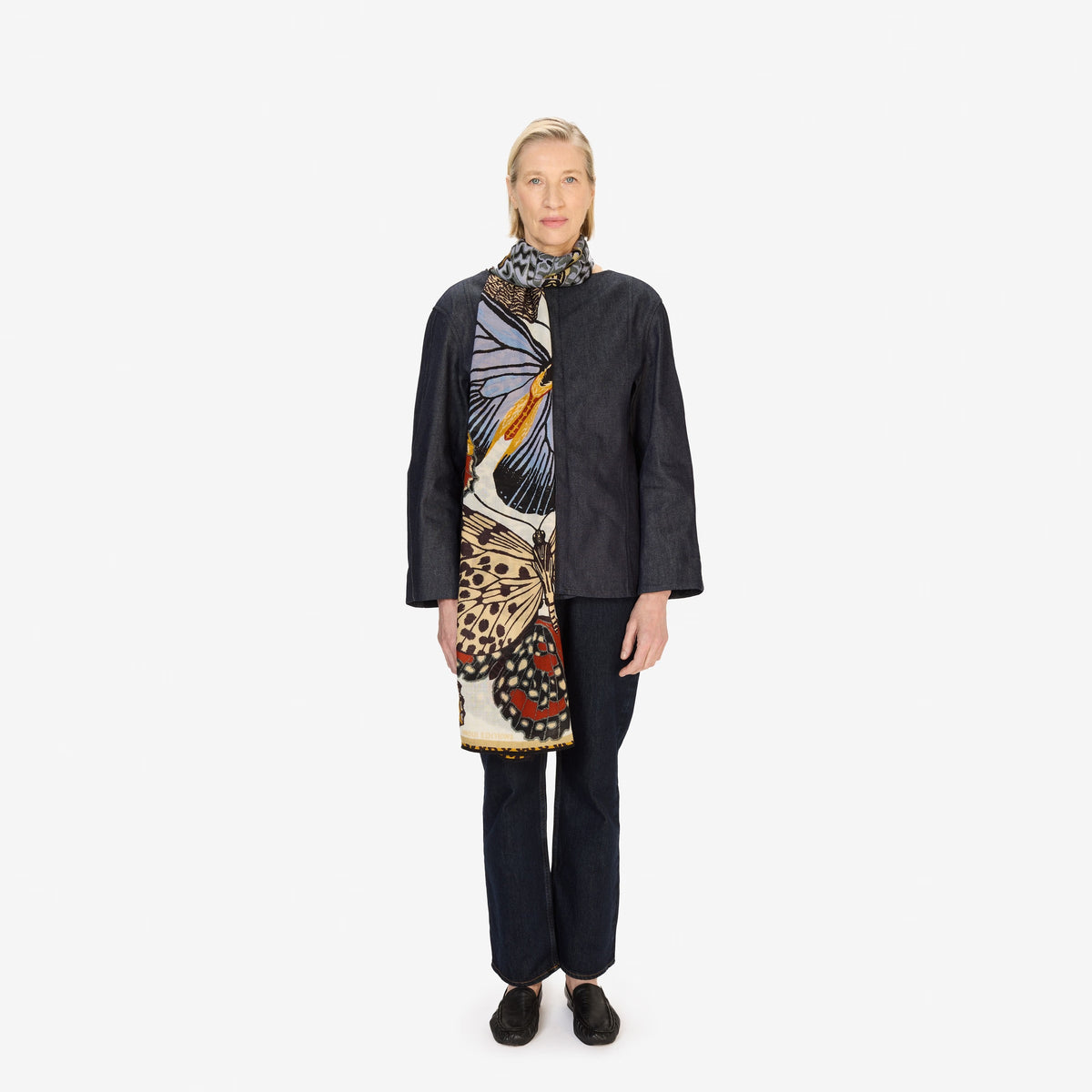 A person dressed in dark attire models the INOUI EDITIONS PAPILLONS (BUTTERFLIES) wool scarf in white by INOUI, featuring a colorful butterfly wing pattern, against a plain white background.