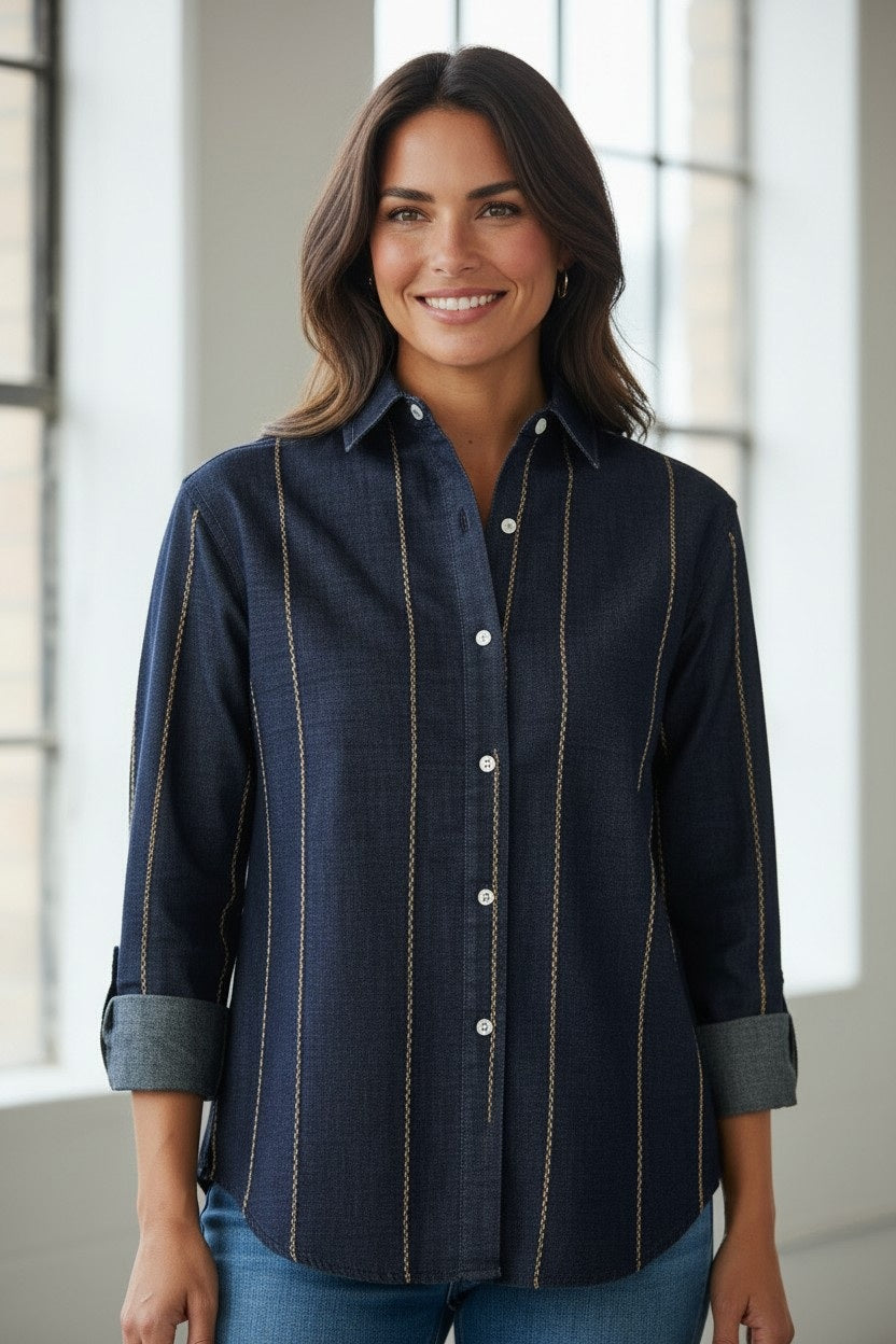 A woman stands indoors by large windows, smiling. She wears the EMILY PHILLIPS Denim with Brown Embroidered Stripe Shirt, featuring long sleeves and button-up front, paired with blue denim jeans.