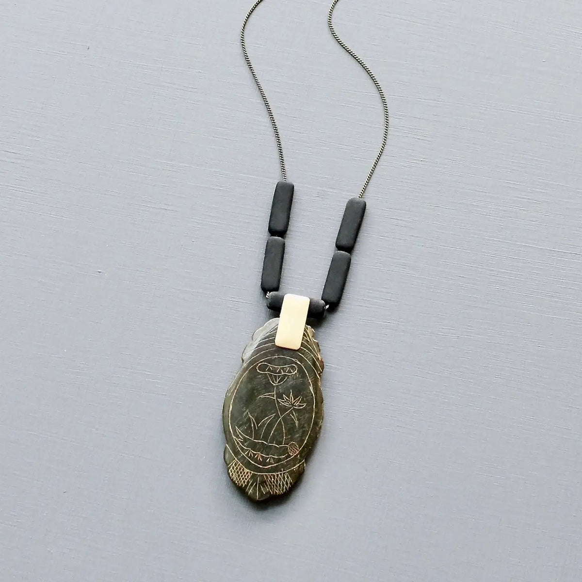 The DAVID AUBREY - CARVED JADE PENDANT NECKLACE by DAVID AUBREY features a 24-inch brass-ox rectangular chain with three black beads and a feather-shaped pendant engraved with an owl and plant design, displayed on a light grey background.