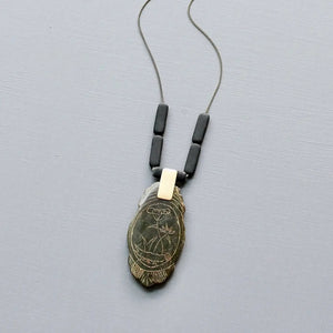 The DAVID AUBREY - CARVED JADE PENDANT NECKLACE by DAVID AUBREY features a 24-inch brass-ox rectangular chain with three black beads and a feather-shaped pendant engraved with an owl and plant design, displayed on a light grey background.