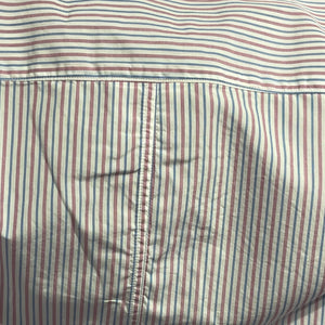 A man with short gray hair and a beard sits on a park bench wearing the FRANK & EILEEN Finbar men's button-up shirt in pink and blue multi stripe, blue jeans, and white sneakers. Behind him, people walk along a flower-lined path among trees.
