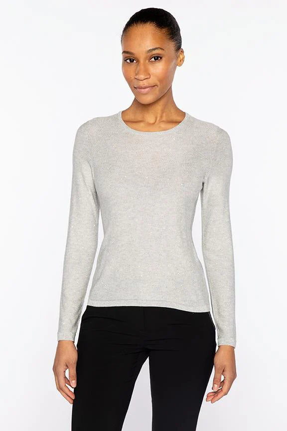 A woman stands facing forward, smiling softly with her hands by her sides, wearing the KINROSS CASHMERE Sequin Crew Neck Sweater in light gray and black pants against a plain white background.