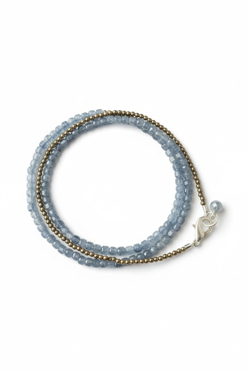 The ANN LIGHTFOOT - SILVERY BLUE KYANITE TRIPLE WRAP BRACELET by ANN LIGHTFOOT is a coiled triple wrap bracelet adorned with small blue and gold beads, finished with a silver clasp and accented by a single blue bead charm near the clasp. Displayed on a white background, its design channels the elegance of kyanite, seamlessly merging modern style with timeless appeal.