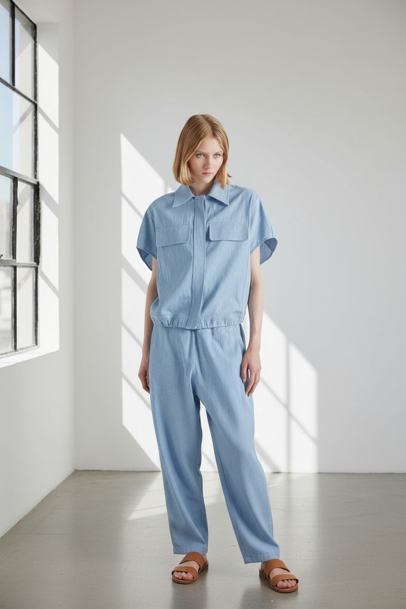A person with chin-length light brown hair stands with their back to the camera, wearing the MEIMEIJ DRAWSTRING COTTON TOP in chambray blue. The loose fit and plain white background highlight MEIMEIJ's modern Italian aesthetic.