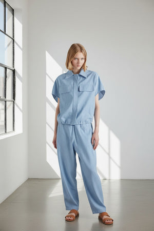 A person with chin-length light brown hair stands with their back to the camera, wearing the MEIMEIJ DRAWSTRING COTTON TOP in chambray blue. The loose fit and plain white background highlight MEIMEIJ's modern Italian aesthetic.