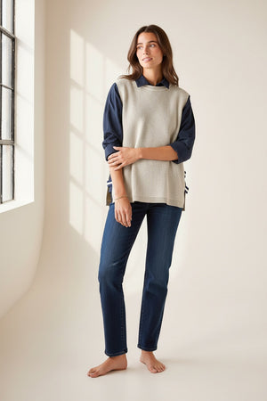 A woman with long brown hair stands sideways against a light background, looking at the camera, wearing the MERSEA - CAMDEN VEST IN DESERT PALM WITH STRIPED TAB by MERSEA over a dark collared shirt and blue jeans.