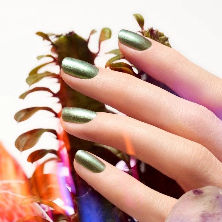 A hand with metallic green nails holds a bottle of MANUCURIST - GREEN™ NAIL POLISH IN GROOVE from MANUCURIST INC, perfectly matching the light green shirt in the background.