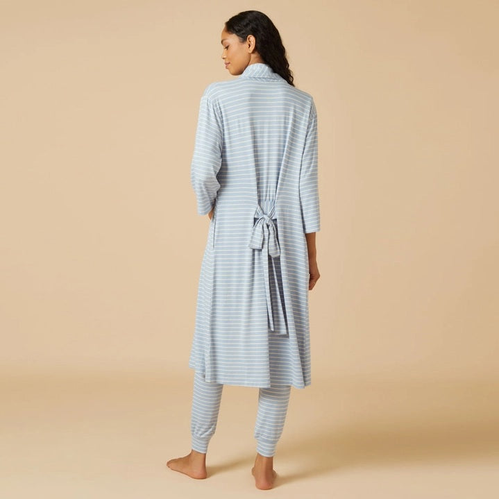Wearing light blue and white striped pajamas, a person stands barefoot against a beige background, draped in the SOFTIES Dream Shawl Collar Robe in Sky Blue Stripe. This lightweight robe from SOFTIES adds an extra layer of silky smooth comfort to the cozy look.
