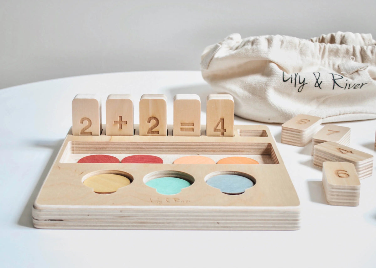 The "LITTLE NUMBERS GAME SET" by LILY &amp; RIVER is a wooden educational toy influenced by Montessori principles. It features number blocks and colored disks on a white surface. The blocks display a simple math equation (2 + 2 = 4), functioning like a children's calculator. In the background, there is a light-colored fabric bag with the text "Lily &amp; River" partially visible.
