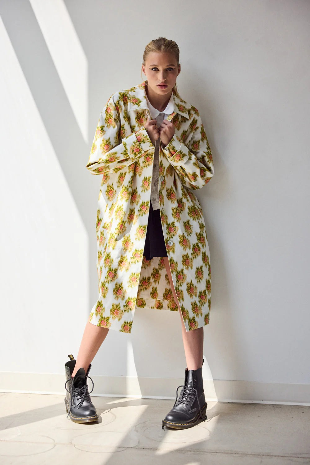 A person stands on grass before green bushes, wearing HOPE FOR FLOWERS' pink Warped Ikat coat over a white shirt and matching shorts with black sandals, looking to the side and highlighting the brand's commitment to ethical fashion.