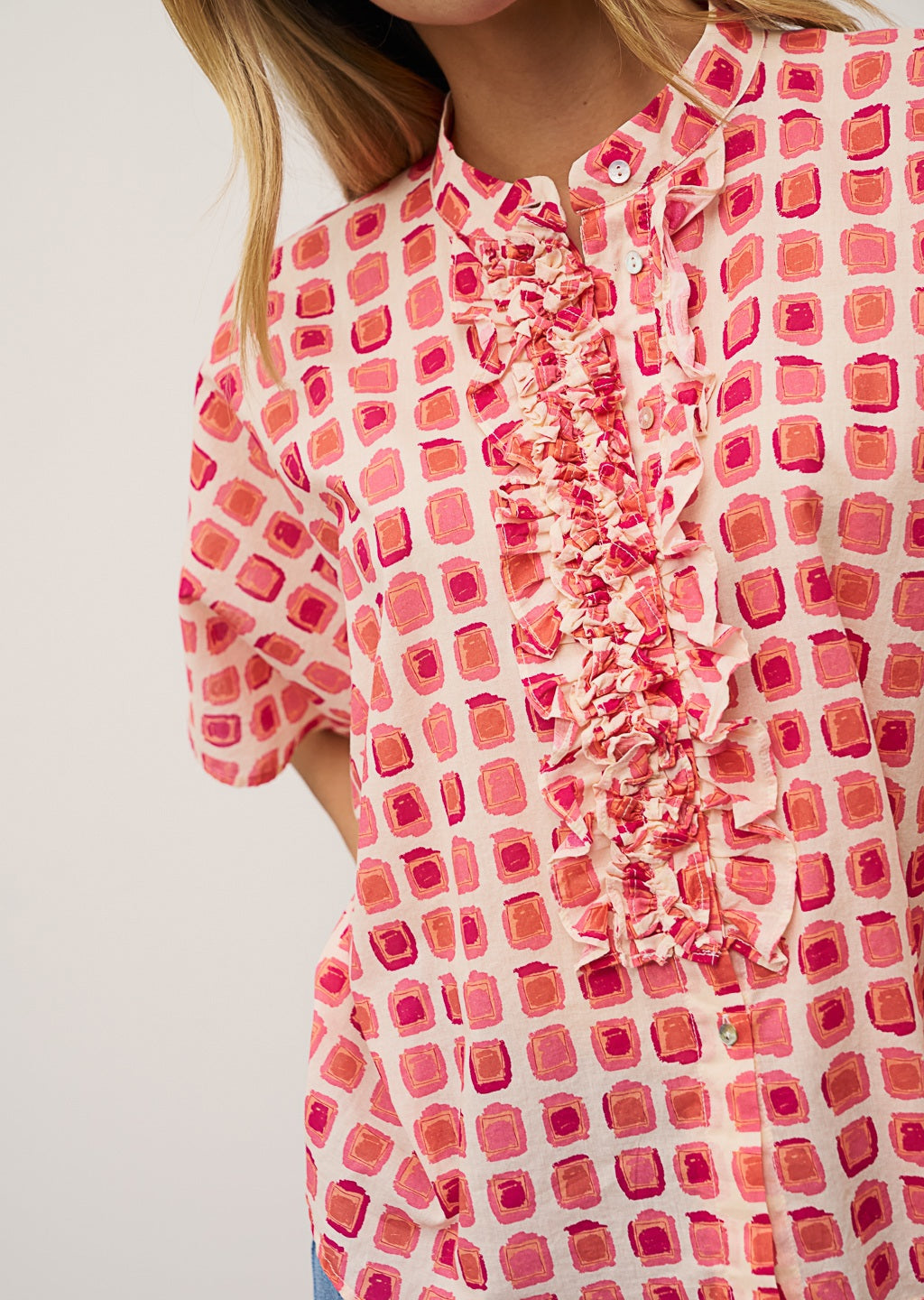 A woman wears the DAISY BLOUSE IN PINK PRINT by 0039 ITALY, featuring pink and red square patterns with ruffles, paired with light blue jeans against a plain light background.