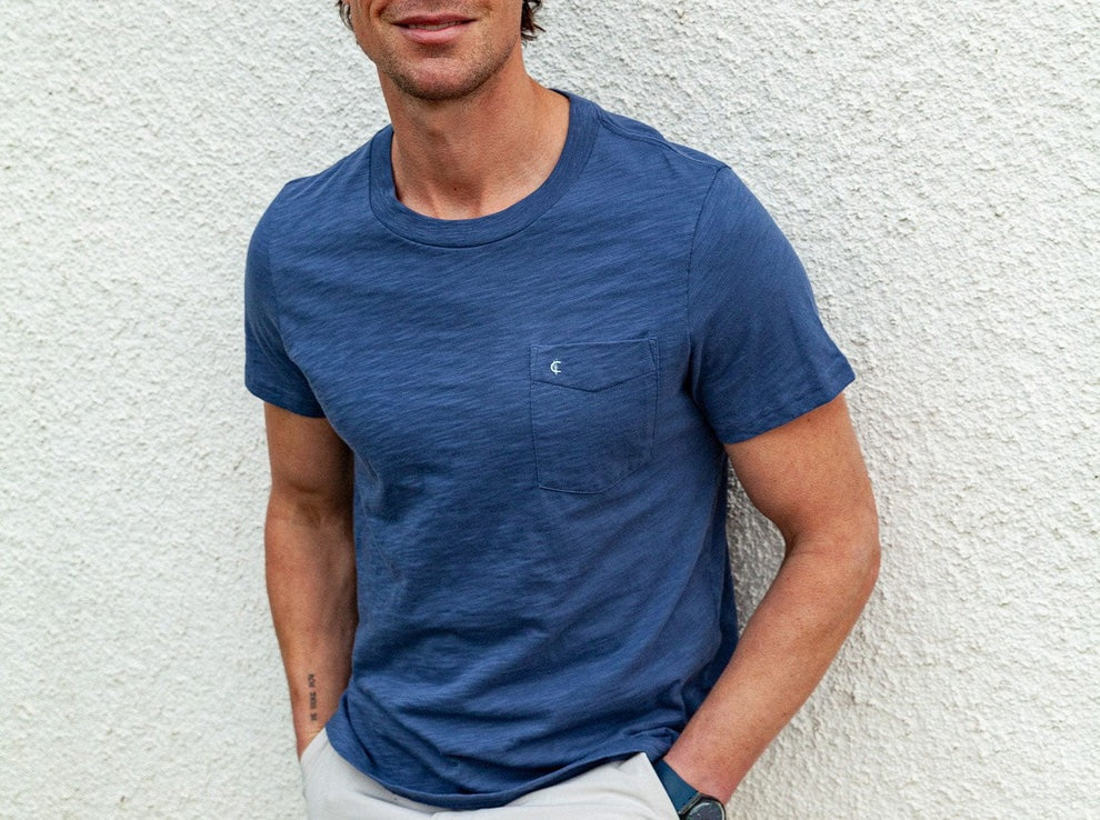 A man is wearing the CRIQUET Slub Cotton Short Sleeve Pocket T-Shirt in Vintage Indigo by CRIQUET, featuring a subtle textured finish and a small chest pocket. He pairs it with light-colored pants and a watch, standing against a white background. The classic fit of the t-shirt complements his relaxed style. His face is partially visible, and he is smiling.