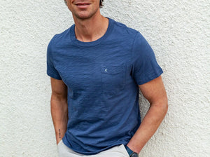 A man is wearing the CRIQUET Slub Cotton Short Sleeve Pocket T-Shirt in Vintage Indigo by CRIQUET, featuring a subtle textured finish and a small chest pocket. He pairs it with light-colored pants and a watch, standing against a white background. The classic fit of the t-shirt complements his relaxed style. His face is partially visible, and he is smiling.