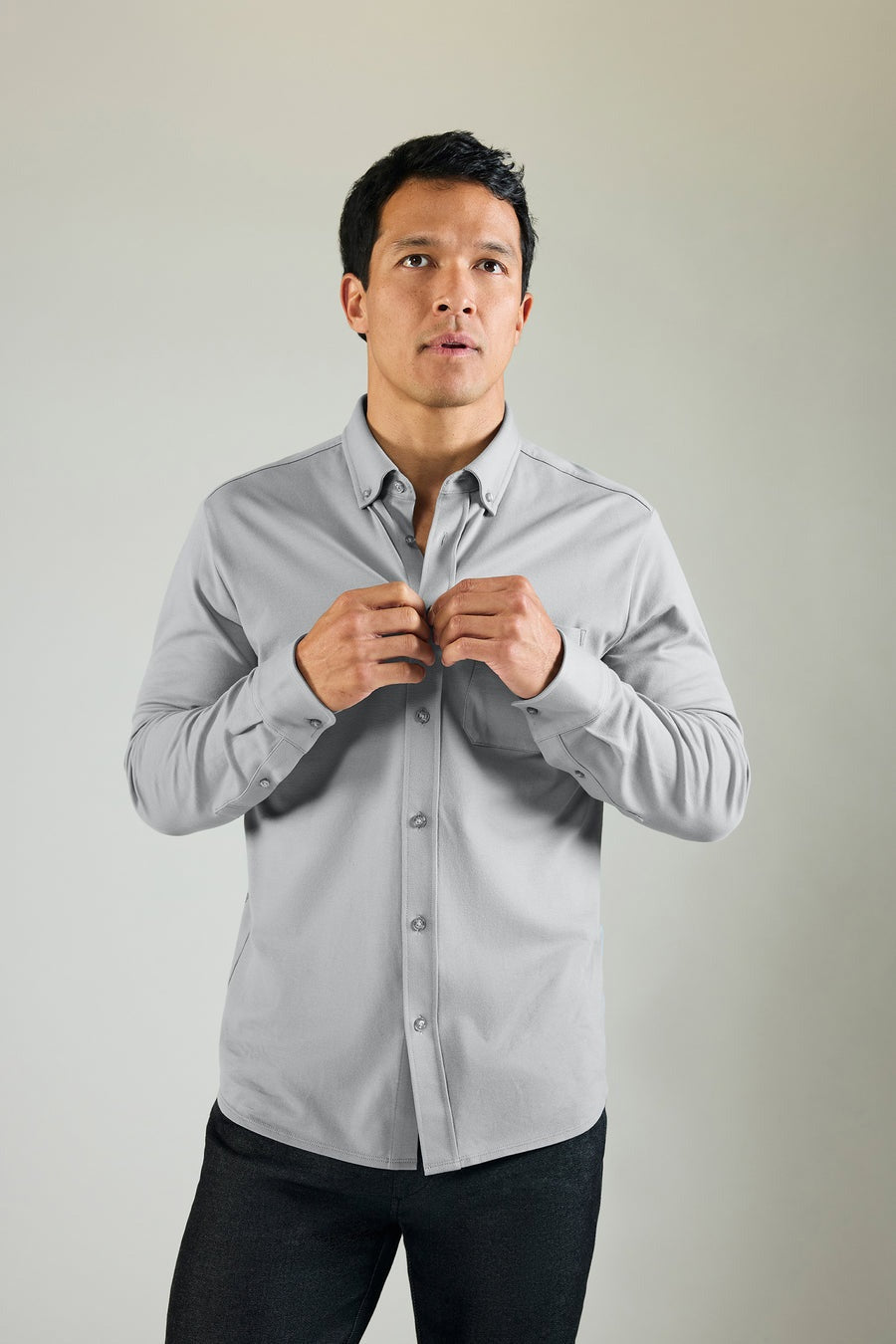 A man faces forward against a plain background, buttoning up a Monument-colored MACK WELDON Silver Pique Long Sleeve Button Up Shirt. He looks slightly upward with a neutral expression and pairs the shirt with dark pants.