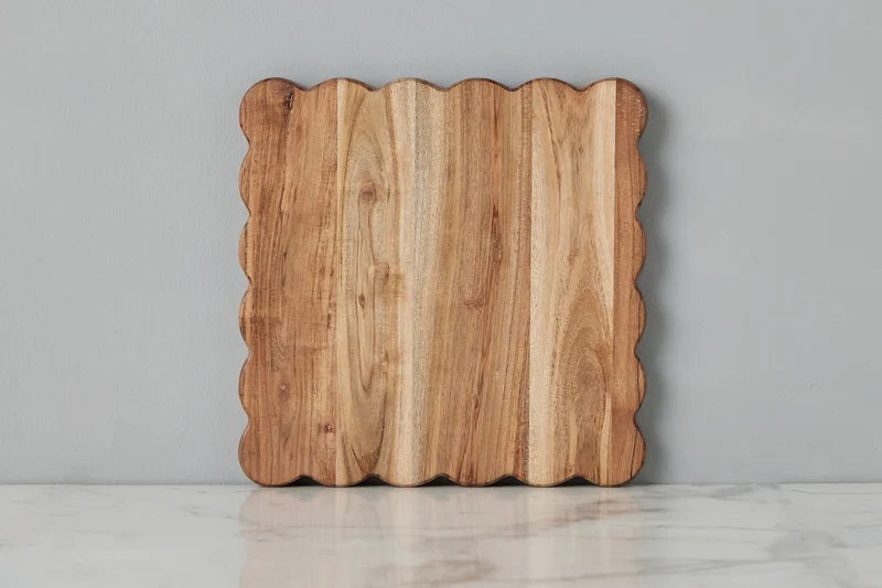 The ETUHOME Natural Square Scalloped Cutting Board, Medium, crafted from durable acacia wood, stands upright against a light gray wall on a white marble surface.