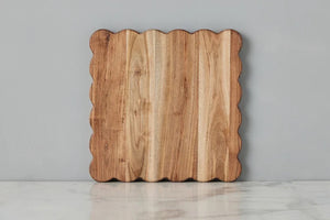 The ETUHOME Natural Square Scalloped Cutting Board, Medium, crafted from durable acacia wood, stands upright against a light gray wall on a white marble surface.