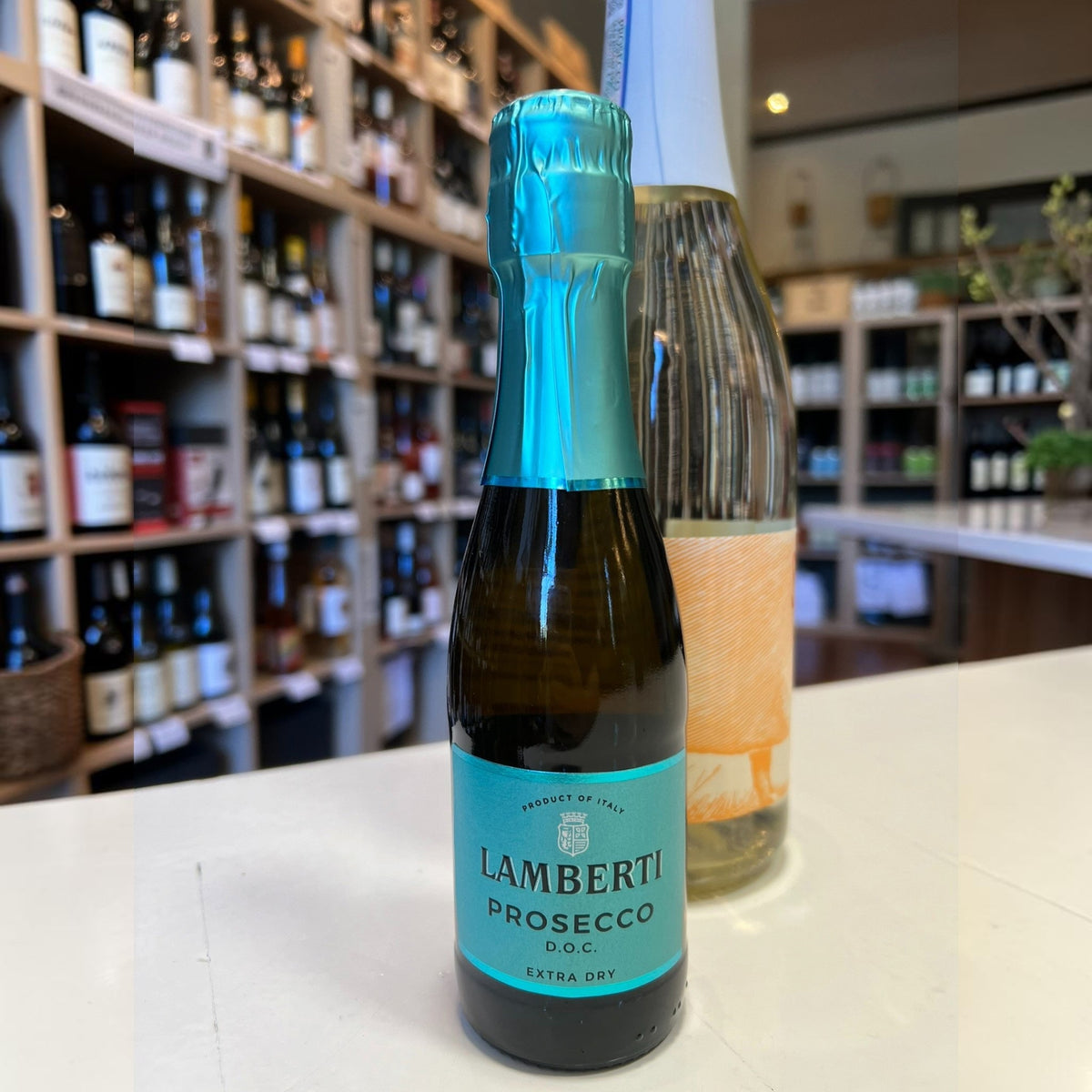 A mini bottle of LAMBERTI PROSECCO 187ML from Veneto, Italy, with a bright blue foil top and matching label, standing on a counter in a wine shop.