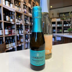 A mini bottle of LAMBERTI PROSECCO 187ML from Veneto, Italy, with a bright blue foil top and matching label, standing on a counter in a wine shop.
