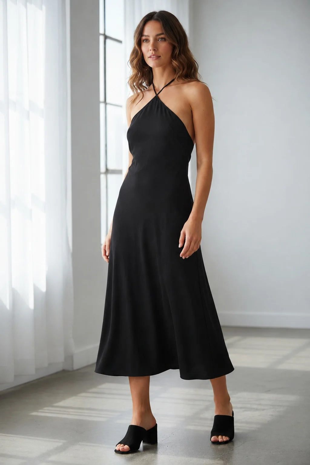 A woman wearing a black midi-length halter dress and black open-toe mules.