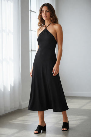A woman faces forward in the SEVENTY JERSEY DRESS, a sleeveless black style with crisscross neckline straps, paired with black open-toe heeled sandals—ideal for summer parties—set against a plain light background.