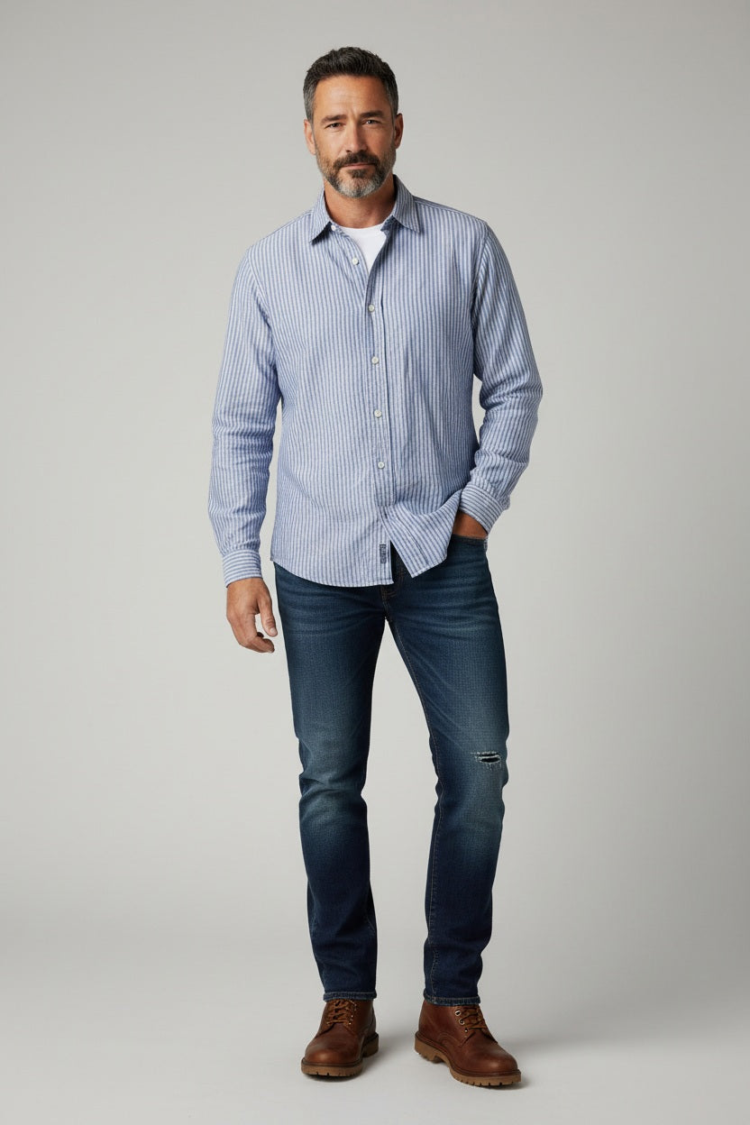 A man in a FRANK &amp; EILEEN Finbar Men's Button Up Shirt in Blue Stripes, blue jeans, and white sneakers walks confidently down a sunny, tree-lined cobblestone street.