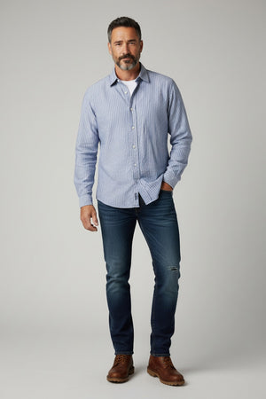 A man in a FRANK & EILEEN Finbar Men's Button Up Shirt in Blue Stripes, blue jeans, and white sneakers walks confidently down a sunny, tree-lined cobblestone street.