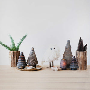 A minimalist winter scene featuring the CREATIVE COOP STONEWARE STAR TEALIGHT HOLDER, stoneware and ceramic pine trees, wooden log vases with pine branches, a fluffy white owl figurine, all arranged on a light wooden table against a plain white wall.