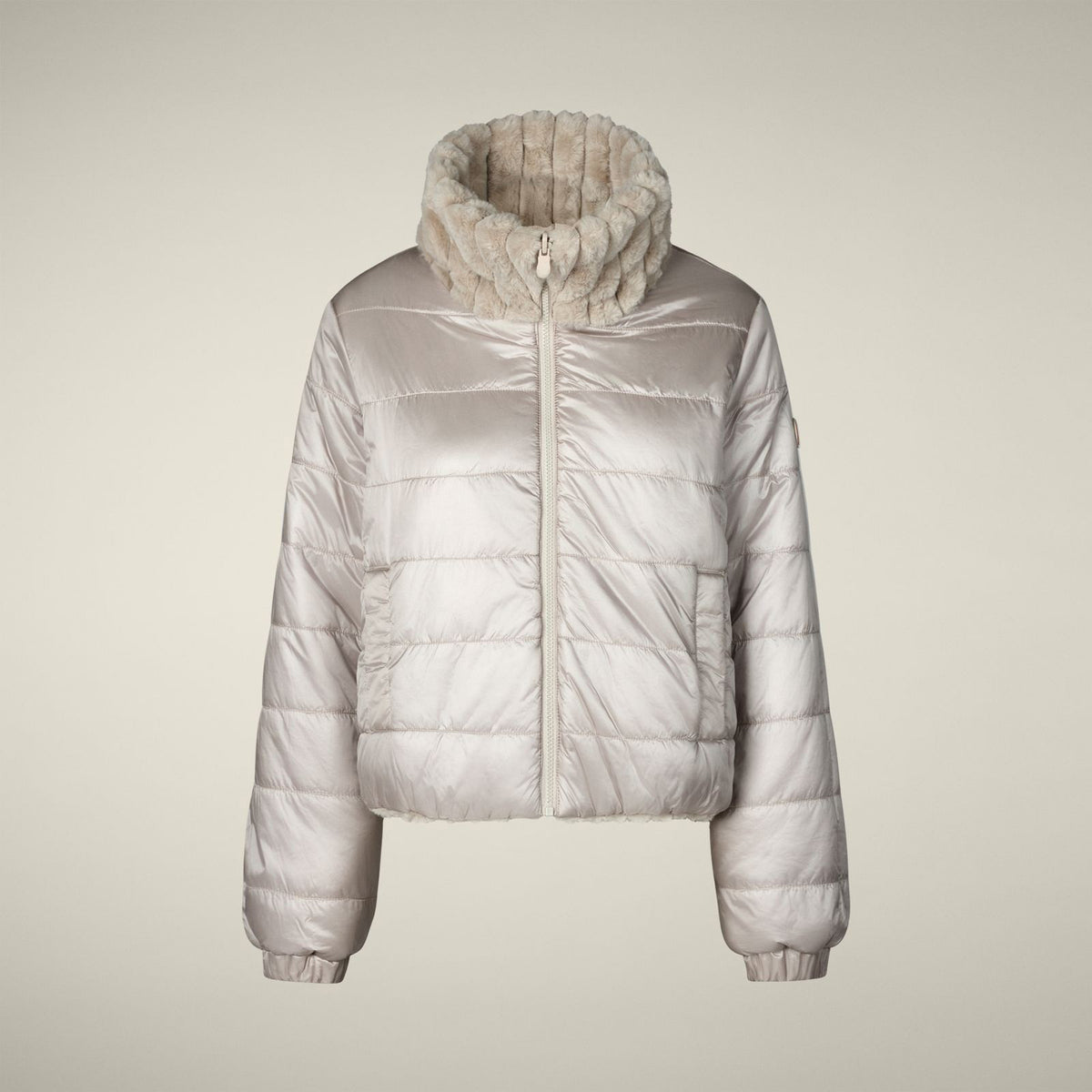 A woman models the SAVE THE DUCK - ELINA REVERSIBLE JACKET in beige fur, styled with a white skirt, orange tights, burgundy boots, and a matching clutch, standing against a plain light background.