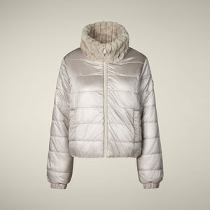 A woman models the SAVE THE DUCK - ELINA REVERSIBLE JACKET in beige fur, styled with a white skirt, orange tights, burgundy boots, and a matching clutch, standing against a plain light background.