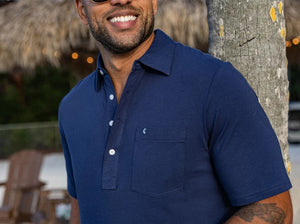 A smiling man in sunglasses stands outside by a wooden building, holding a drink and wearing the CRIQUET Top-Shelf Players Shirt in Navy. Sunlight and greenery are visible in the background.