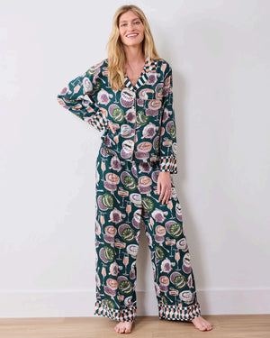 A woman stands barefoot on a wooden floor against a white wall, wearing the PRINTFRESH - Champagne & Caviar Eco Satin Soho Set in Peacock Feather by PRINTFRESH, a matching dark loungewear set adorned with colorful floral and fruit motifs and finished with checkered cuffs.