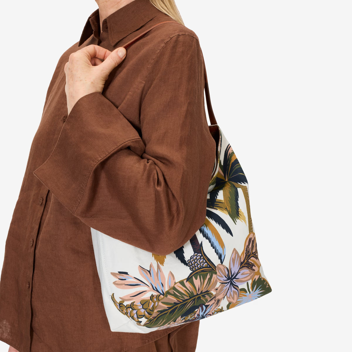 The INOUI EDITIONS - HAWAII CAMPUS CANVAS BAG highlights a tropical landscape in earthy tones with palm trees and foliage, offering a painterly vintage charm. Its slouchy design and single leather strap elegantly combine style and function against a plain white background.