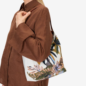 The INOUI EDITIONS - HAWAII CAMPUS CANVAS BAG highlights a tropical landscape in earthy tones with palm trees and foliage, offering a painterly vintage charm. Its slouchy design and single leather strap elegantly combine style and function against a plain white background.