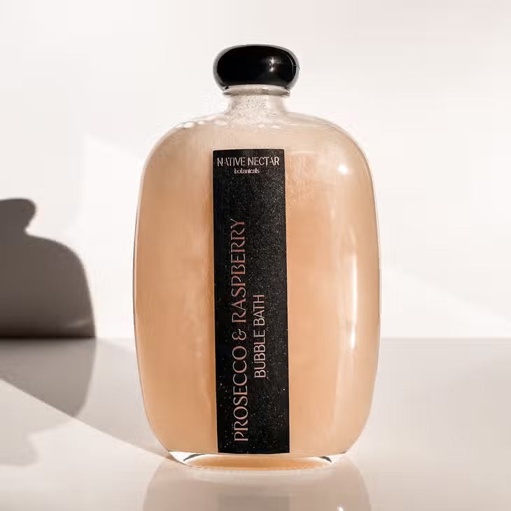 Beneath soft lighting, a clear bottle branded with "NATIVE NECTAR's PROSECCO AND RASPBERRY BUBBLE BATH" displays its hydrating pink contents. Topped with a round cap, it sits gracefully on a reflective surface, offering a luxurious bath experience enriched with natural ingredients.