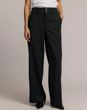A person wears HINSON WU's KELLY TRAVELER'S TWILL PANT in dark green with front pockets and gold buttons, styled with a white top and black pointed-toe shoes. Photo is cropped from shoulders down, highlighting this travel-ready look.