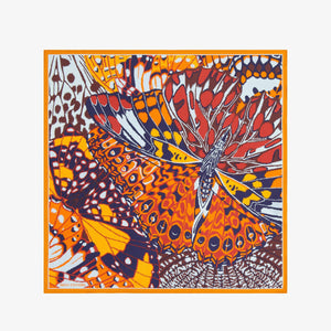 A person in a dark grey knit sweater wears an INOUI EDITIONS PAPILLONS Orange Silk Scarf, featuring bold abstract butterfly designs and “INOUI EDITIONS” on the border, neatly tied around their neck.
