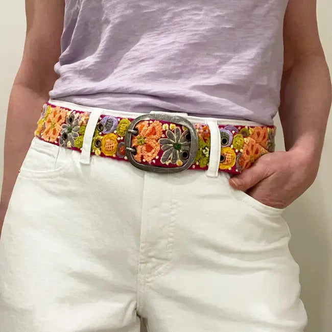 The JENNY KRAUSS WILDFLOWERS EMBROIDERED WOOL BELT features hand-embroidered floral patterns in orange, yellow, green, purple, and white on a peach background, crafted in Peru with a silver buckle and shown coiled on a white surface.
